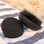 PUR HAIR Detangler Brush, in schwarz, liegt auf gestreiftem Handtuch, am Strand.