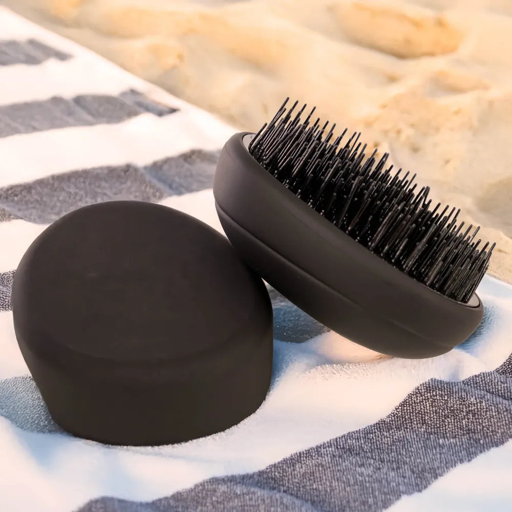 PUR HAIR Detangler Brush, in schwarz, liegt auf gestreiftem Handtuch, am Strand.