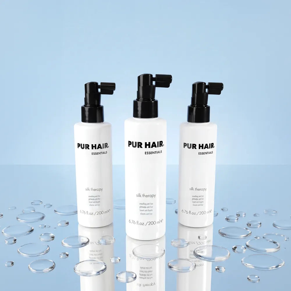 Drei Flaschen PUR HAIR Silk Therapy Anti-Frizz-Spray auf hellblauem Hintergrund mit Wassertropfen.