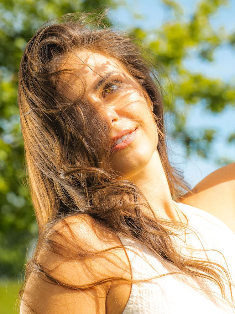 Frau mit langem, glänzendem braunem Haar im Sonnenlicht vor grünem Hintergrund.