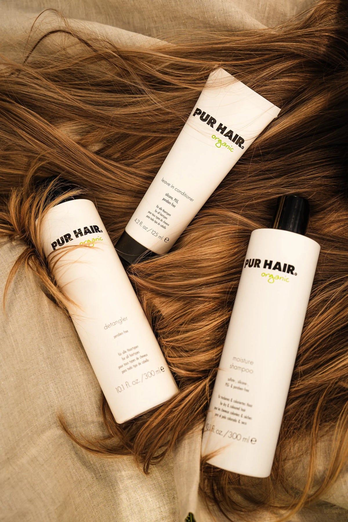 Drei weiße PUR HAIR Produkte liegen auf braunem Haar und braunem Leinenstoff-Untergrund.