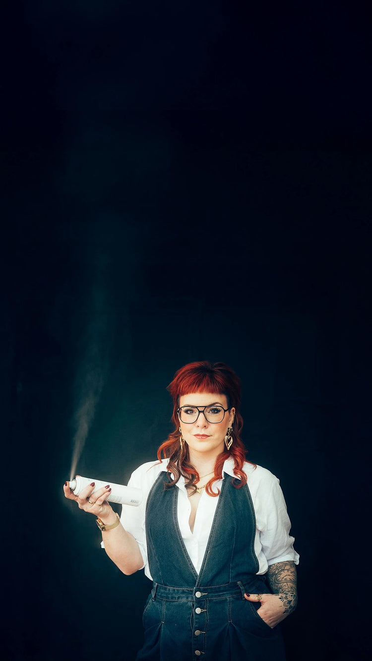 Friseurin mit rotem Haar und Brille hält ein PUR HAIR Stylingprodukt und sprüht feinen Sprühnebel.