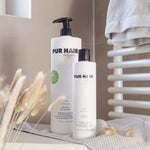 Weiße Flaschen PUR HAIR Organic Magic Treatment 1000 ml und 125 ml, auf grauem Tuch mit Blumen.