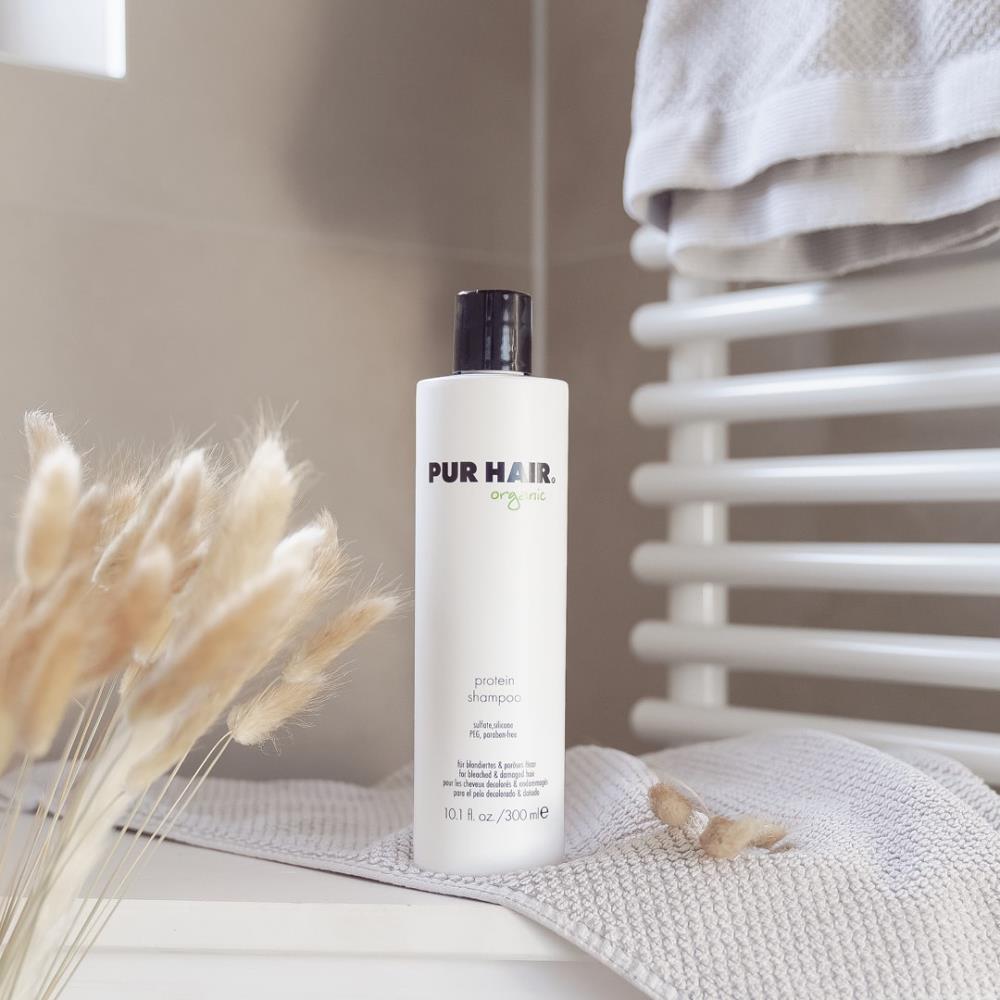 Weiße Flasche PUR HAIR Organic Protein Shampoo, 300 ml, auf grauem Handtuch im Badezimmer platziert.