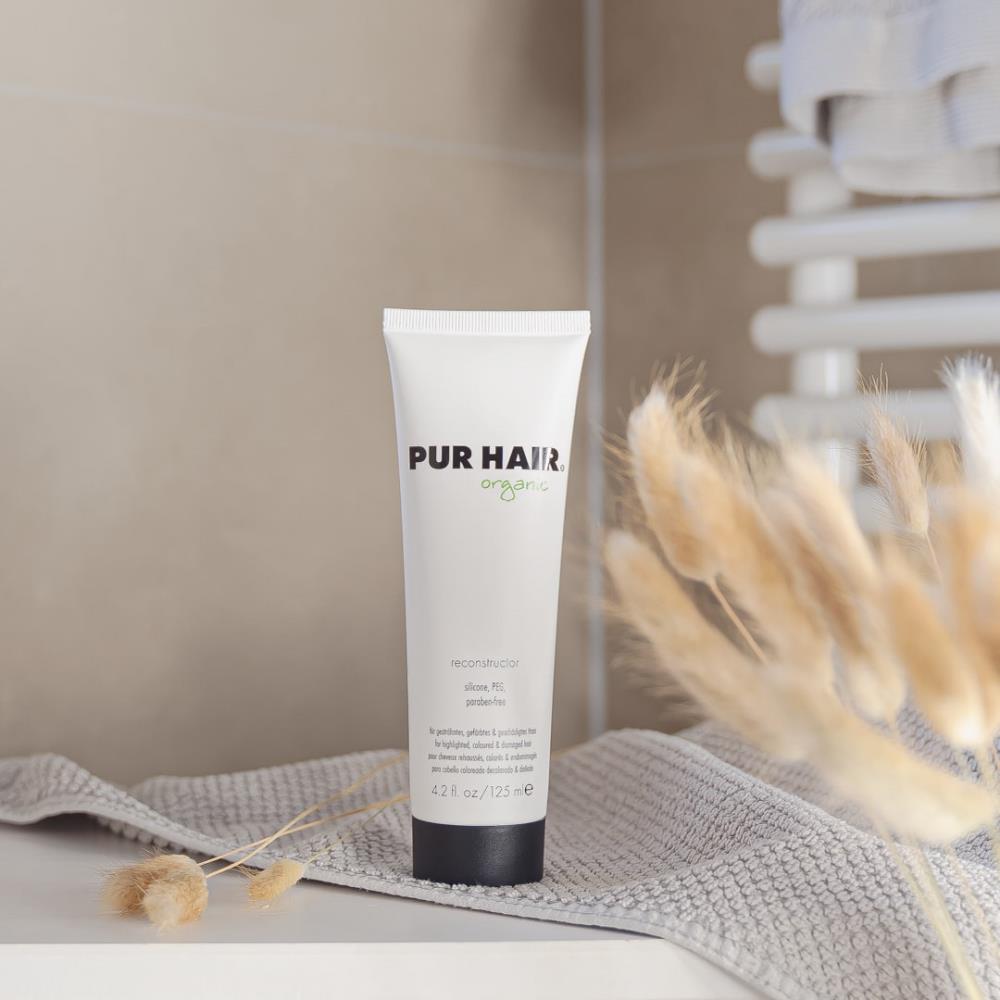 Weiße 125-ml-Tube PUR HAIR Organic Reconstructor mit schwarzem Deckel, aufgestellt neben Vase.